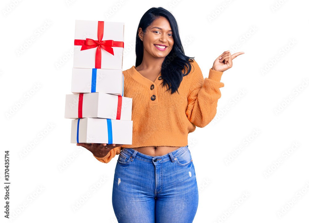 Hispanic woman with long hair holding gifts smiling happy pointing with hand and finger to the side Hispanic woman with long hair holding gifts smiling happy pointing with hand and finger to the side