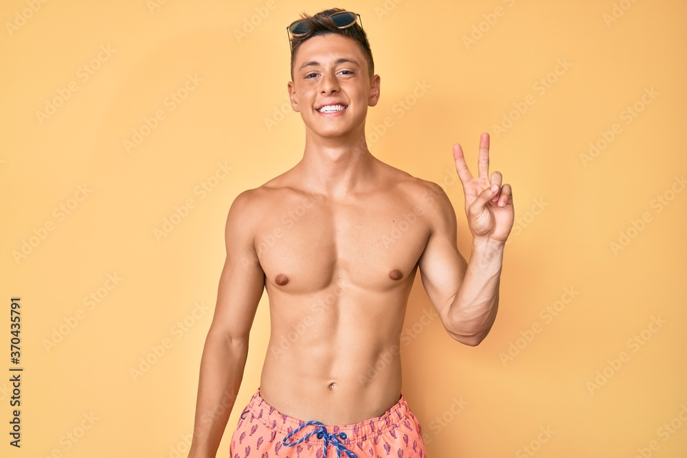 Young hispanic boy wearing swimwear shirtless smiling with happy face winking at the camera doing victory sign. number two.