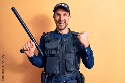 Handsome policeman wearing uniform and bulletprof holding baton over yellow background pointing thumb up to the side smiling happy with open mouth photo