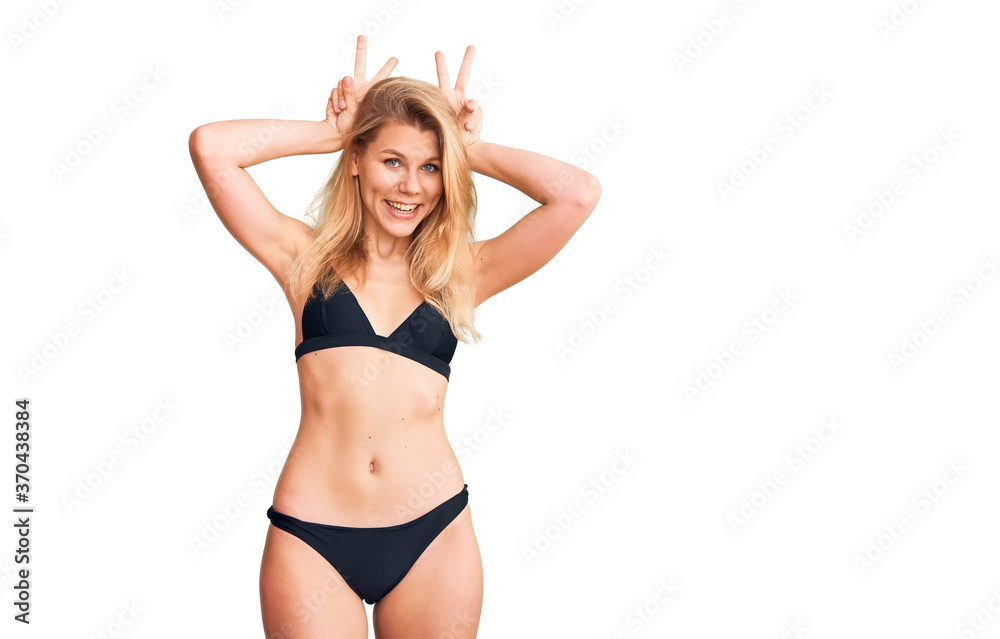 Young beautiful blonde woman wearing bikini posing funny and crazy with fingers on head as bunny ears, smiling cheerful