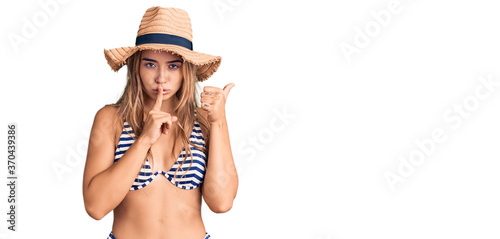 Young beautiful blonde woman wearing bikini and hat asking to be quiet with finger on lips pointing with hand to the side. silence and secret concept. photo