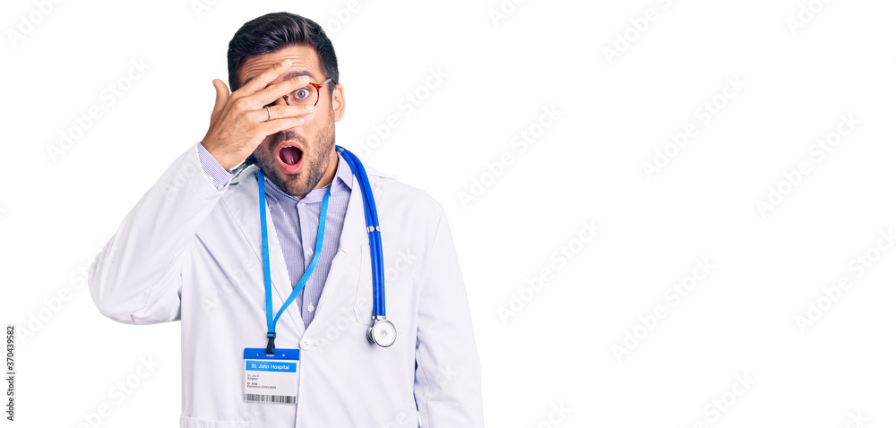 Young hispanic man wearing doctor uniform and stethoscope peeking in shock covering face and eyes with hand, looking through fingers with embarrassed expression. Young hispanic man wearing doctor uniform and stethoscope peeking in shock covering face and eyes with hand, looking through fingers with embarrassed expression.