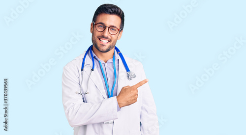 Young hispanic man wearing doctor uniform and stethoscope cheerful with a smile on face pointing with hand and finger up to the side with happy and natural expression photo