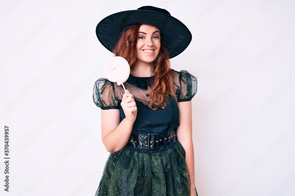 Young beautiful woman wearing witch halloween costume holding lollipop looking positive and happy standing and smiling with a confident smile showing teeth