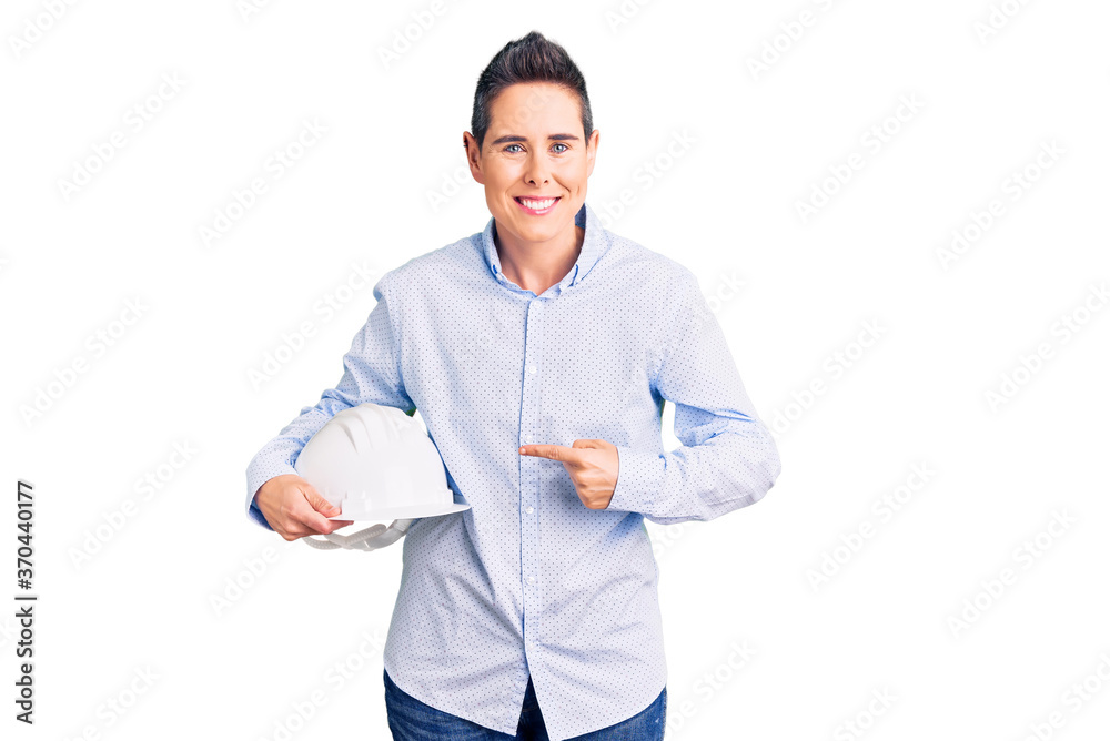 Young woman with short hair holding architect hardhat smiling happy pointing with hand and finger
