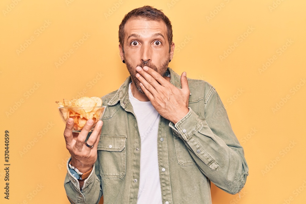 Young handsome man holding potato chip covering mouth with hand, shocked and afraid for mistake. surprised expression Young handsome man holding potato chip covering mouth with hand, shocked and afraid for mistake. surprised expression
