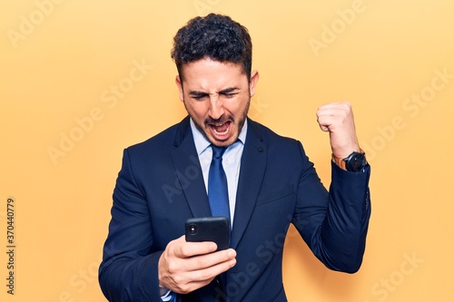 Young hispanic man wearing suit using smartphone screaming proud, celebrating victory and success very excited with raised arms © Krakenimages.com