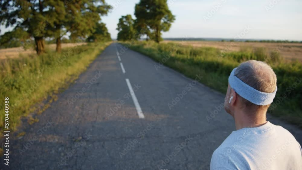 Adult man runs on empty road with portable headphones and blue headband for sportsman. Male runner summer uniform. Active acations. Leisure activity. Physical education. Nature freshness. Healthcare. Adult man runs on empty road with portable headphones and blue headband for sportsman. Male runner summer uniform. Active acations. Leisure activity. Physical education. Nature freshness. Healthcare.