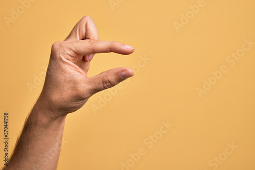 Hand of caucasian young man showing fingers over isolated yellow background picking and taking invisible thing, holding object with fingers showing space