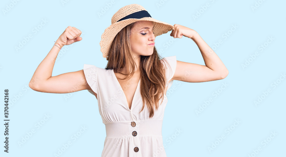 Young beautiful blonde woman wearing summer dress and hat showing arms muscles smiling proud. fitness concept. Young beautiful blonde woman wearing summer dress and hat showing arms muscles smiling proud. fitness concept.