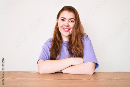 Young redhead woman wearing casual clothes sitting on the table happy face smiling with crossed arms looking at the camera. positive person.