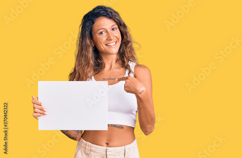 Young hispanic woman with tattoo holding blank empty paper pointing finger to one self smiling happy and proud