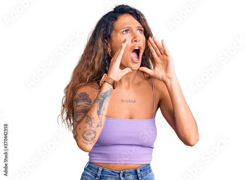 Young hispanic woman with tattoo wearing casual clothes shouting angry out loud with hands over mouth