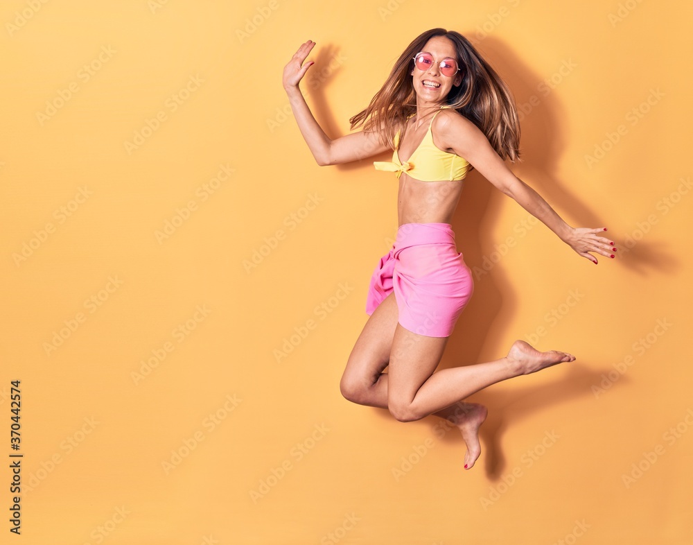 Young beautiful girl wearing bikini and sunglasses smiling happy. Jumping with smile on face over isolated yellow background Young beautiful girl wearing bikini and sunglasses smiling happy. Jumping with smile on face over isolated yellow background