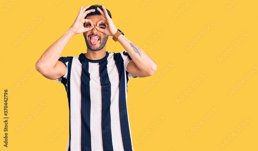 Young handsome hispanic man wearing striped tshirt doing ok gesture like binoculars sticking tongue out, eyes looking through fingers. crazy expression.