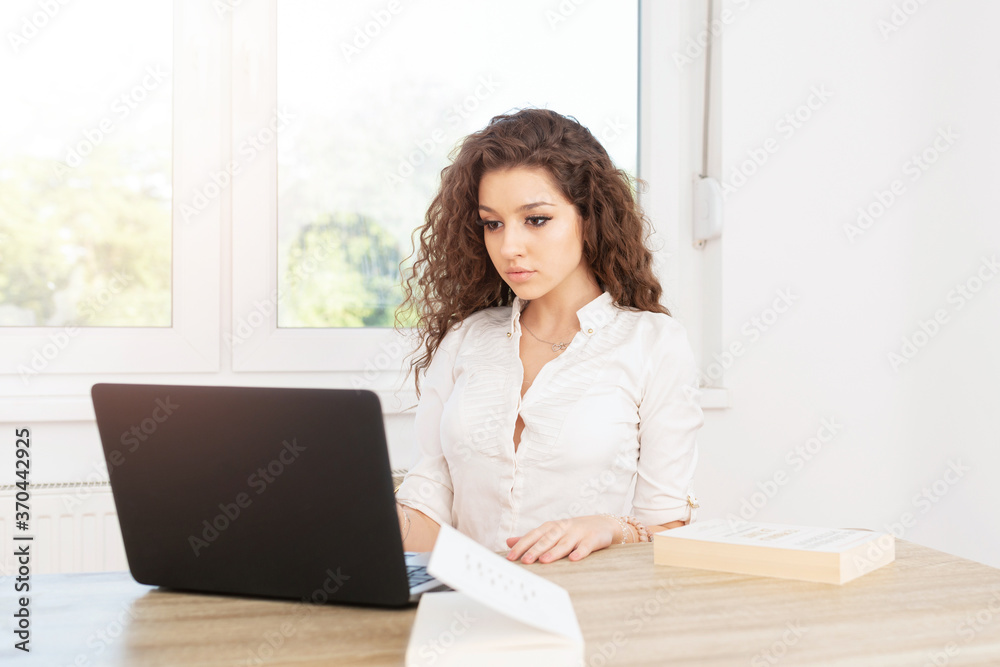 Elegant businesswoman with curly brown hair in white shirt sitting in office by the window and looking at laptop. Elegant businesswoman with curly brown hair in white shirt sitting in office by the window and looking at laptop.