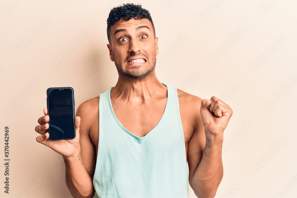 Young hispanic man holding smartphone showing screen screaming proud, celebrating victory and success very excited with raised arm Young hispanic man holding smartphone showing screen screaming proud, celebrating victory and success very excited with raised arm