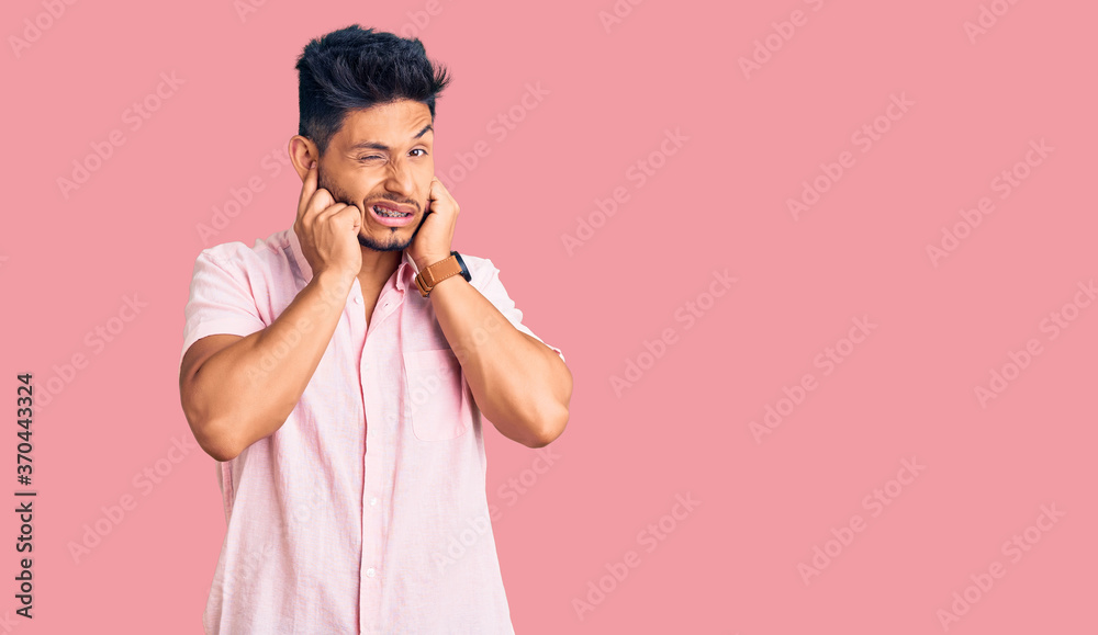 Handsome latin american young man wearing casual summer shirt covering ears with fingers with annoyed expression for the noise of loud music. deaf concept. Handsome latin american young man wearing casual summer shirt covering ears with fingers with annoyed expression for the noise of loud music. deaf concept.