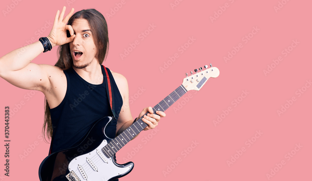 Young adult man with long hair playing electric guitar smiling happy doing ok sign with hand on eye looking through fingers Young adult man with long hair playing electric guitar smiling happy doing ok sign with hand on eye looking through fingers