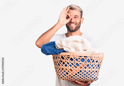 Young handsome blond man holding laundry basket with clothes smiling happy doing ok sign with hand on eye looking through fingers