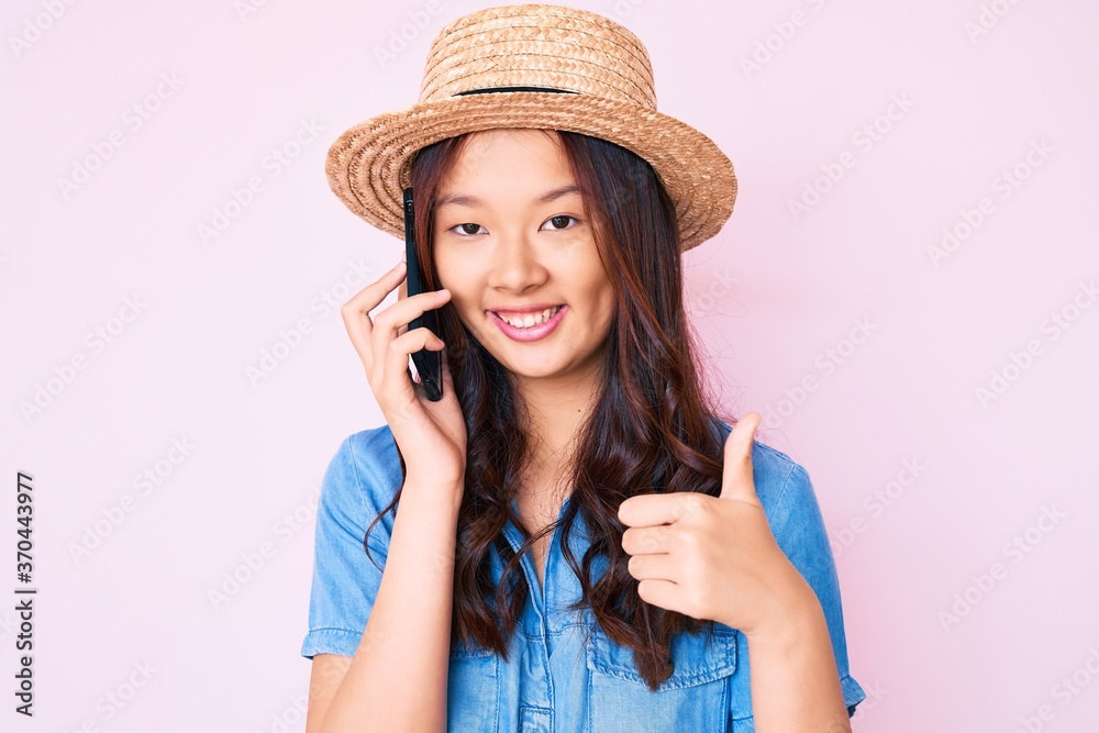 Young beautiful chinese girl wearing summer hat having conversation talking on the smartphone smiling happy and positive, thumb up doing excellent and approval sign Young beautiful chinese girl wearing summer hat having conversation talking on the smartphone smiling happy and positive, thumb up doing excellent and approval sign