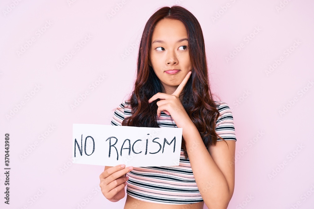 Young beautiful chinese girl holding no racism banner serious face thinking about question with hand on chin, thoughtful about confusing idea Young beautiful chinese girl holding no racism banner serious face thinking about question with hand on chin, thoughtful about confusing idea