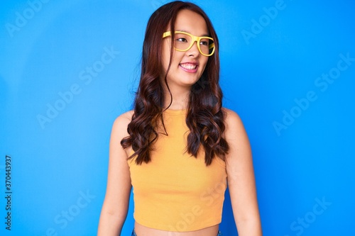 Young beautiful chinese girl wearing casual clothes and glasses looking away to side with smile on face, natural expression. laughing confident.