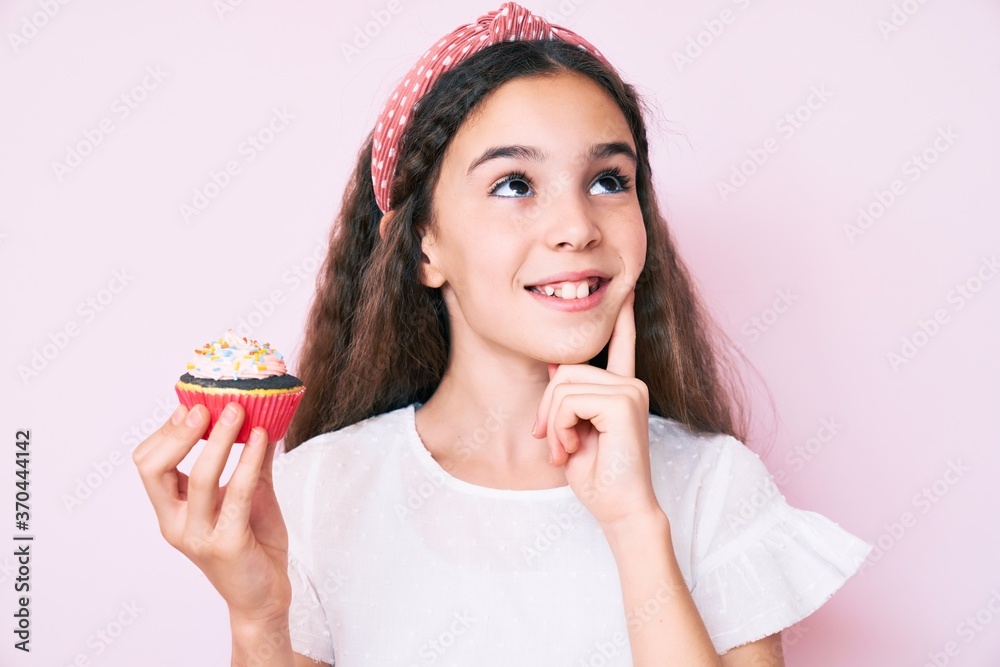 Cute hispanic child girl holding cupcake serious face thinking about question with hand on chin, thoughtful about confusing idea Cute hispanic child girl holding cupcake serious face thinking about question with hand on chin, thoughtful about confusing idea