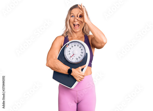 Middle age fit blonde woman wearing sports clothes holding weighing machine smiling happy doing ok sign with hand on eye looking through fingers