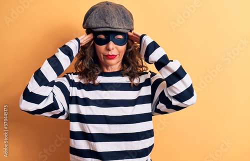 Middle age brunette burglar woman wearing mask and cap standing over yellow background with hand on head, headache because stress. Suffering migraine.