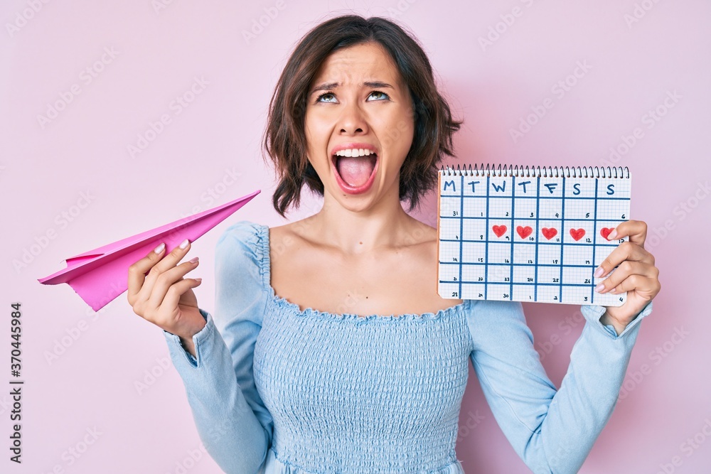 Young beautiful woman holding heart calendar and paper airplane angry
