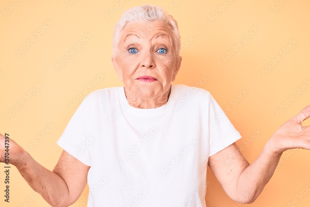 Senior beautiful woman with blue eyes and grey hair wearing classic white tshirt over yellow background clueless and confused expression with arms and hands raised. doubt concept. Senior beautiful woman with blue eyes and grey hair wearing classic white tshirt over yellow background clueless and confused expression with arms and hands raised. doubt concept.