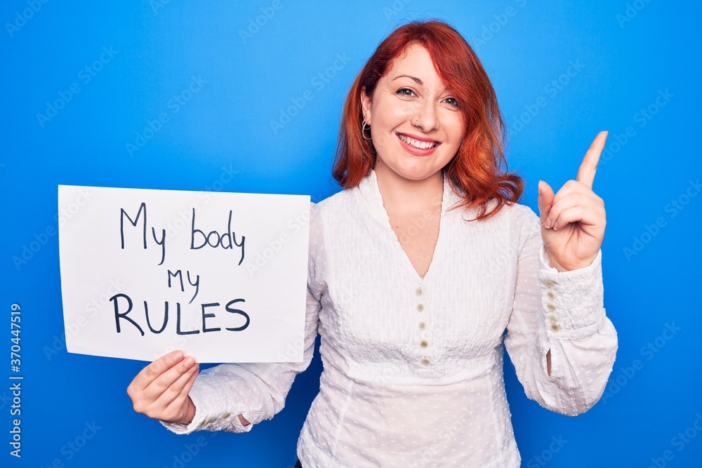 Young redhead woman asking for women rights holding paper with my body my rules message smiling happy pointing with hand and finger to the side Young redhead woman asking for women rights holding paper with my body my rules message smiling happy pointing with hand and finger to the side