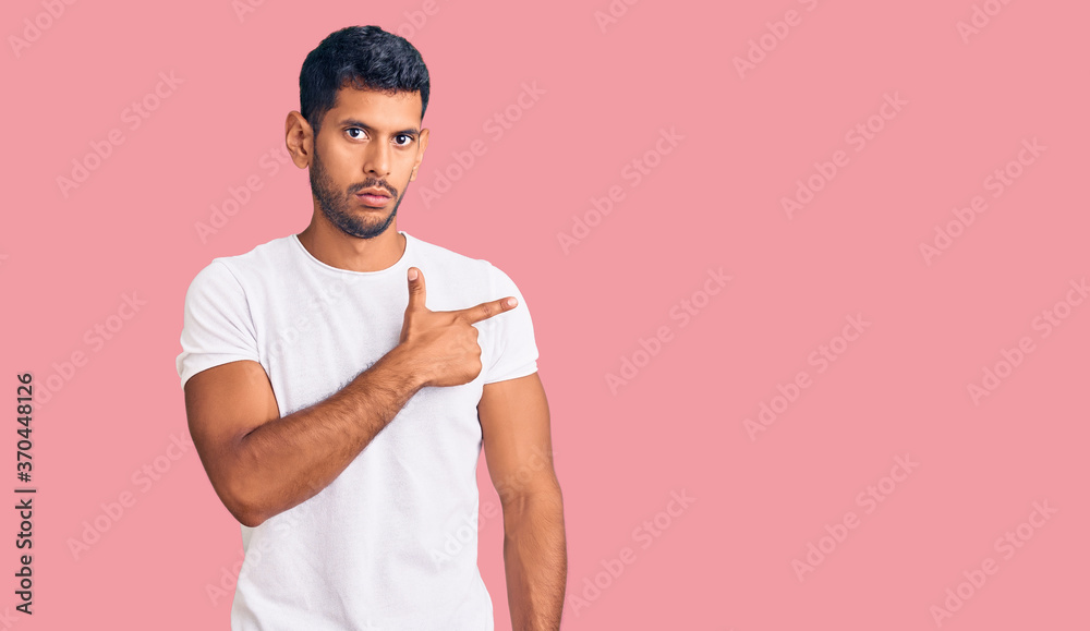 Young latin man wearing casual clothes pointing with hand finger to the side showing advertisement, serious and calm face Young latin man wearing casual clothes pointing with hand finger to the side showing advertisement, serious and calm face