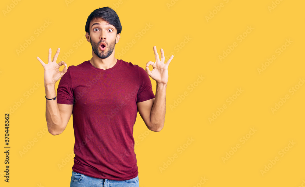 Handsome hispanic man wearing casual clothes looking surprised and shocked doing ok approval symbol with fingers. crazy expression