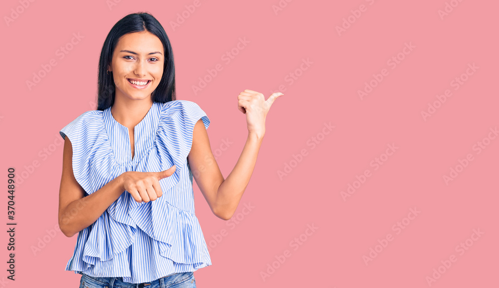 Young beautiful latin girl wearing casual clothes pointing to the back behind with hand and thumbs up, smiling confident Young beautiful latin girl wearing casual clothes pointing to the back behind with hand and thumbs up, smiling confident
