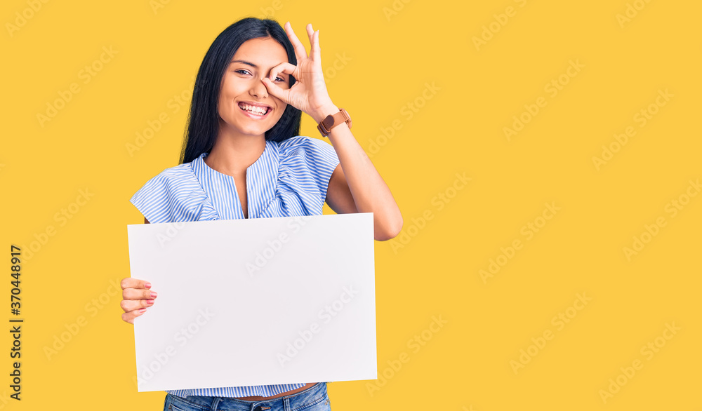 Young beautiful latin girl holding blank empty banner smiling happy doing ok sign with hand on eye looking through fingers Young beautiful latin girl holding blank empty banner smiling happy doing ok sign with hand on eye looking through fingers