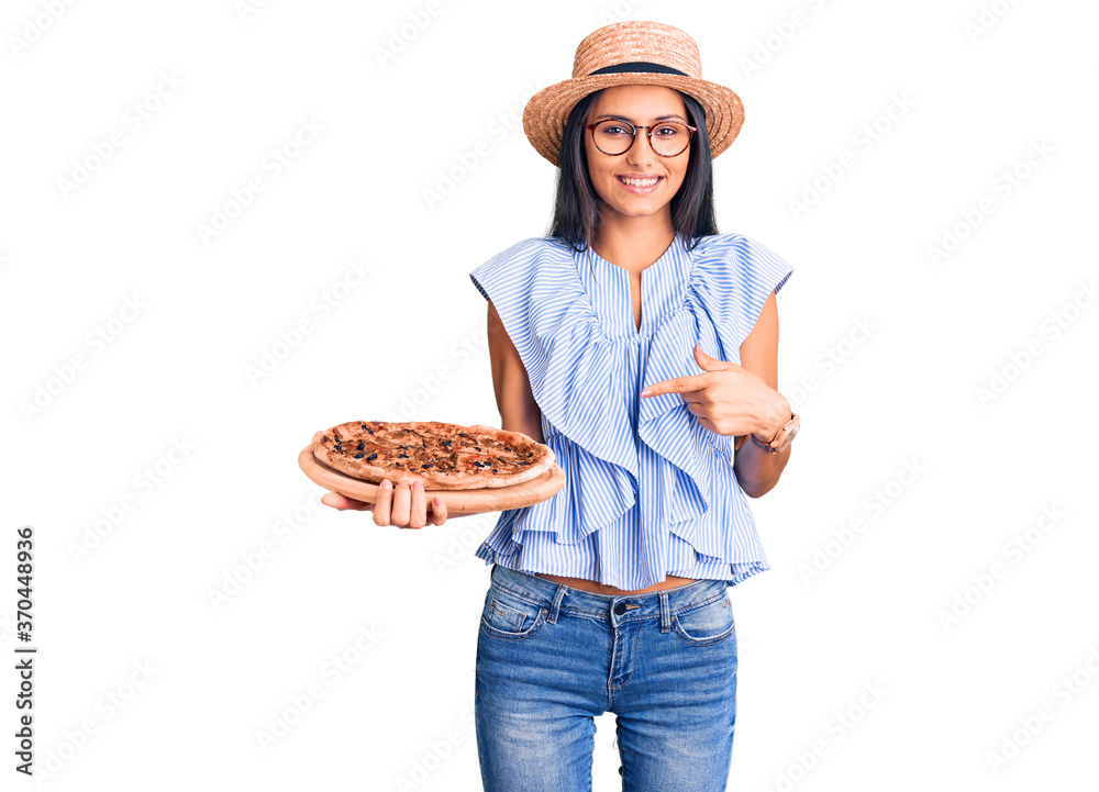 Young beautiful latin girl wearing summer hat and glasses holding pizza smiling happy pointing with hand and finger Young beautiful latin girl wearing summer hat and glasses holding pizza smiling happy pointing with hand and finger