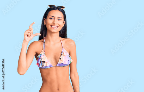 Young beautiful latin girl wearing bikini and sunglasses smiling and confident gesturing with hand doing small size sign with fingers looking and the camera. measure concept.