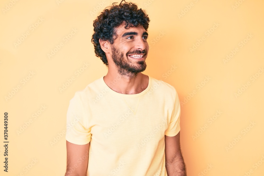 Handsome young man with curly hair and bear wearing casual tshirt over yellow background looking to side, relax profile pose with natural face and confident smile. Handsome young man with curly hair and bear wearing casual tshirt over yellow background looking to side, relax profile pose with natural face and confident smile.