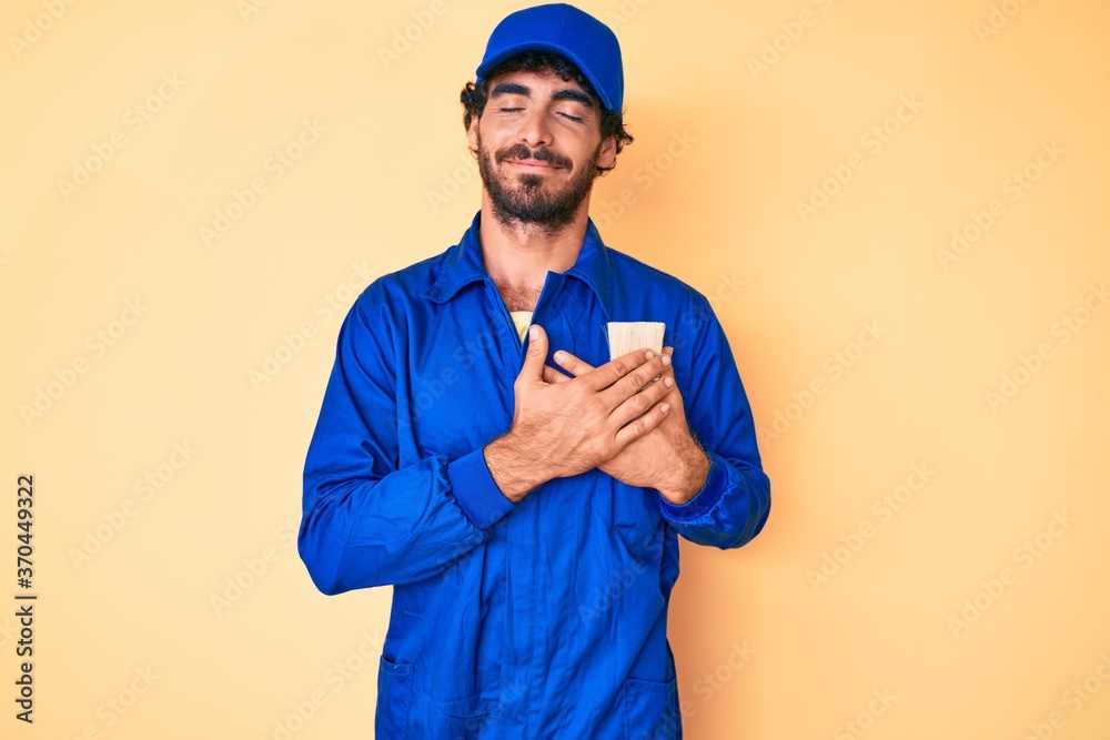 Handsome young man with curly hair and bear wearing builder jumpsuit uniform smiling with hands on chest with closed eyes and grateful gesture on face. health concept.