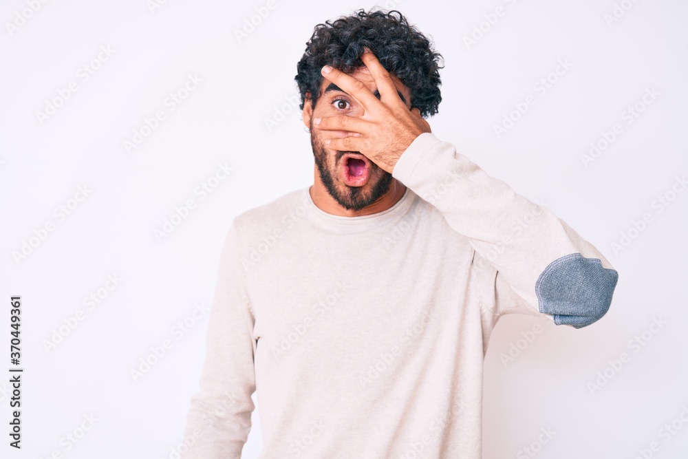 Handsome young man with curly hair and bear wearing casual winter sweater peeking in shock covering face and eyes with hand, looking through fingers afraid Handsome young man with curly hair and bear wearing casual winter sweater peeking in shock covering face and eyes with hand, looking through fingers afraid