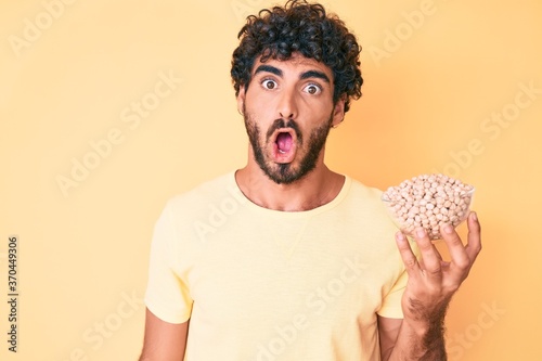 Tableau sur toile Handsome young man with curly hair and bear holding chickpeas bowl scared and am