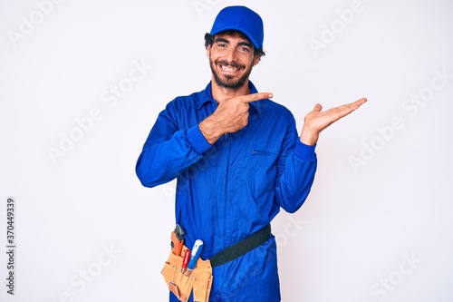 Handsome young man with curly hair and bear weaing handyman uniform amazed and smiling to the camera while presenting with hand and pointing with finger.