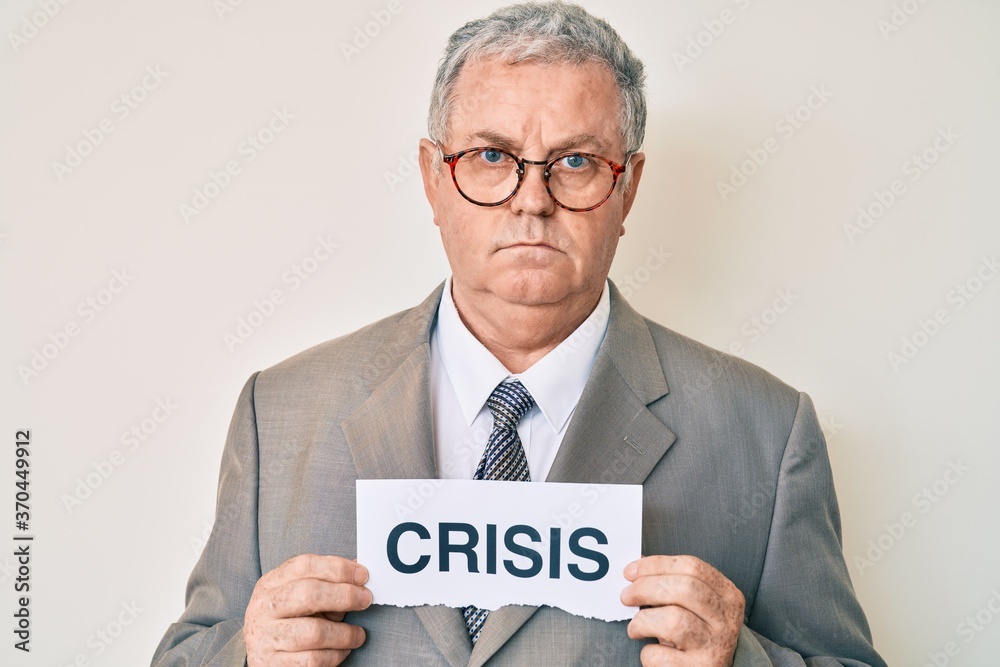 Senior grey-haired man wearing business suit holding crisis paper relaxed with serious expression on face. simple and natural looking at the camera. Senior grey-haired man wearing business suit holding crisis paper relaxed with serious expression on face. simple and natural looking at the camera.