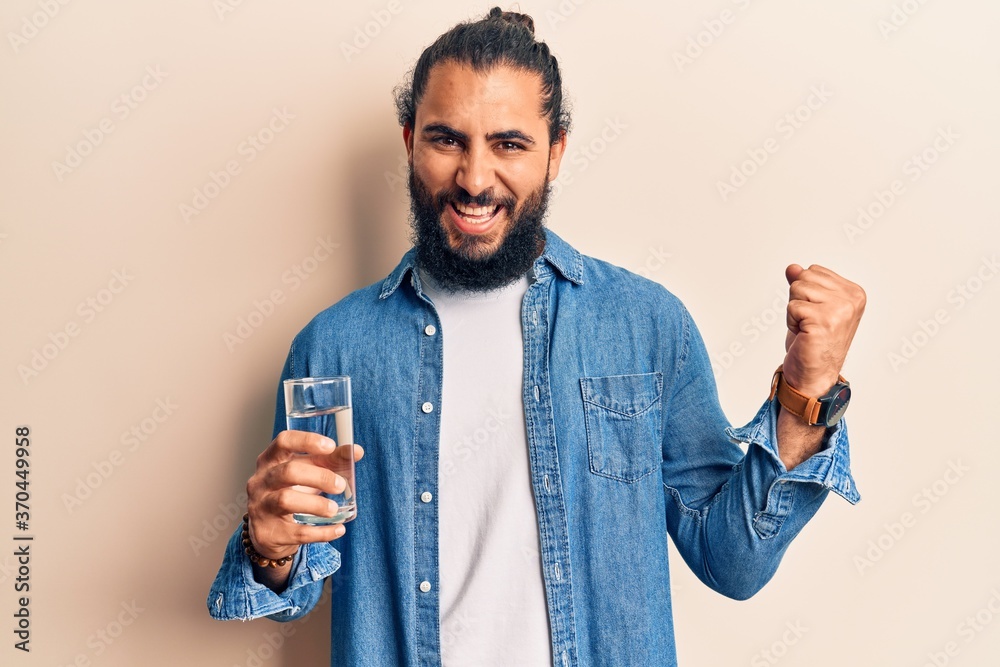 Young arab man drinking glass of water screaming proud, celebrating victory and success very excited with raised arm Young arab man drinking glass of water screaming proud, celebrating victory and success very excited with raised arm