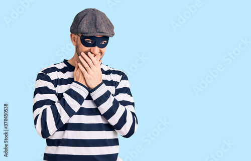 Schilderij op canvas Young handsome man wearing burglar mask laughing and embarrassed giggle covering