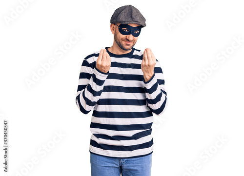 Young handsome man wearing burglar mask doing money gesture with hands, asking for salary payment, millionaire business