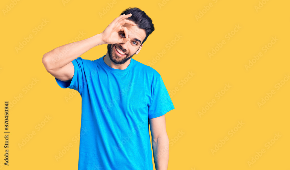 Young handsome man with beard wearing casual t-shirt smiling happy doing ok sign with hand on eye looking through fingers