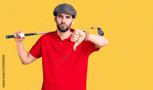 Young handsome man with beard playing golf holding club and ball with angry face, negative sign showing dislike with thumbs down, rejection concept